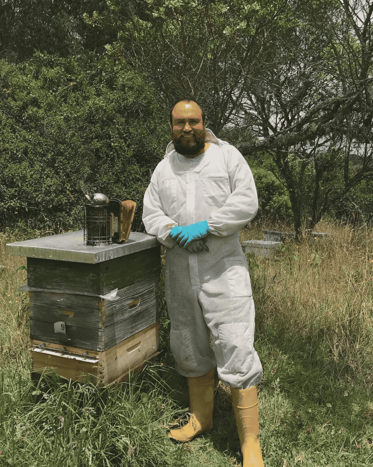 Apicultor con traje protector junto a una colmena, con un ahumador sobre la tapa, en un apiario rodeado de vegetación.